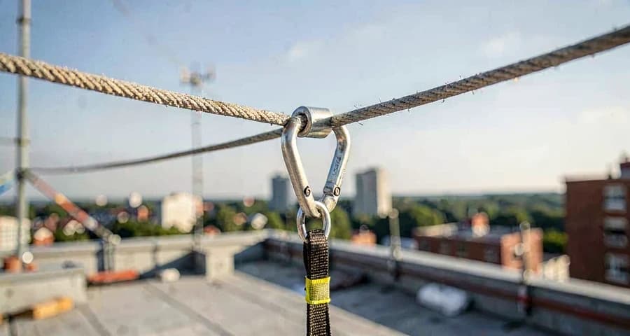Detalhe de mosquetão e talabarte com absorvedor de energia conectados a uma linha de vida em cobertura de obra — Sistema de Proteção Individual Contra Quedas, NR-35